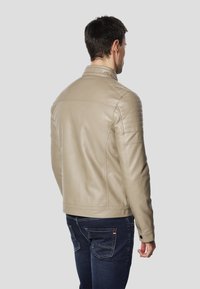 Beżowa skórzana kurtka z prążkowanymi akcentami na ramionach i stojącym kołnierzem, zestawiona z ciemnoniebieskimi dżinsami z widocznymi przeszyciami.