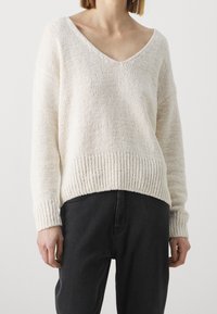 Gebreide trui met een V-hals, gemaakt van crèmekleurig garen, met losse mouwen en een geribbelde onderrand. Gecombineerd met zwarte broek.