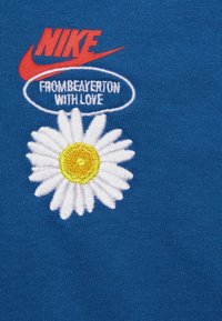 Tissu en coton bleu présentant le logo Nike rouge en haut, une marguerite blanche brodée avec un centre jaune, et le texte "DE BEAVERTON AVEC AMOUR" en blanc.