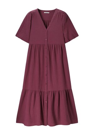 Jeune femme en robe bordeaux fluide et chaussures marron cloutées pose devant un fond beige, la main reposant sur le menton et les jambes visibles à travers la fente.