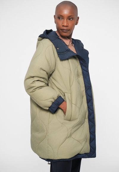 Quilted puffer jacket in olive green with a navy hood and trim, featuring side pockets and a slightly oversized fit. Textured surface detail.