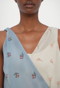 Robe légère avec panneaux bleus et crème, ornée de broderies florales. Le tissu est lisse, avec un design drapé et un nœud sur une épaule.