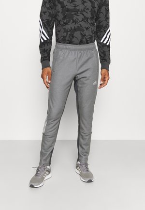 Mężczyzna w szarych spodniach dresowych Adidas z białymi paskami po bokach i szarych butach Adidas, stojący na jednolitym tle.
