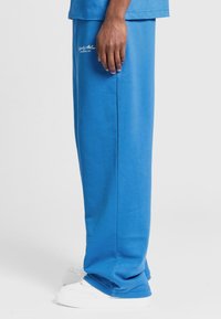 Bl�å bomulds sweatpants med bred benvidde og afslappet pasform, med en sidesøm og hvide sneakers på en ensfarvet baggrund.