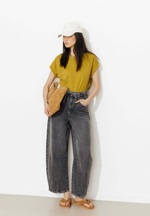 Femme portant une casquette blanche, un t-shirt moutarde, un jean gris ample taille haute, des sandales camel, et tenant un sac souple camel contre un mur blanc.