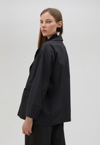 Černá, lehká bunda s přiléhavým střihem, širokými rukávy a límečkem. Má přední kapsu a hladkou, matnou texturu.