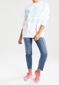 Valkoinen huppari, jossa on mintunvihreä Adidas-logo, pyöreä kaula-aukko ja raglan-hihat. Yhdistetty vaaleansinisiin farkkuihin ja vaaleanpunaisiin lenkkareihin.