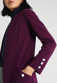 Kobieta w ciemnofioletowej marynarce z srebrnymi guzikami, granatowym golfie, z ręką w kieszeni, pokazana od ramion do połowy uda.