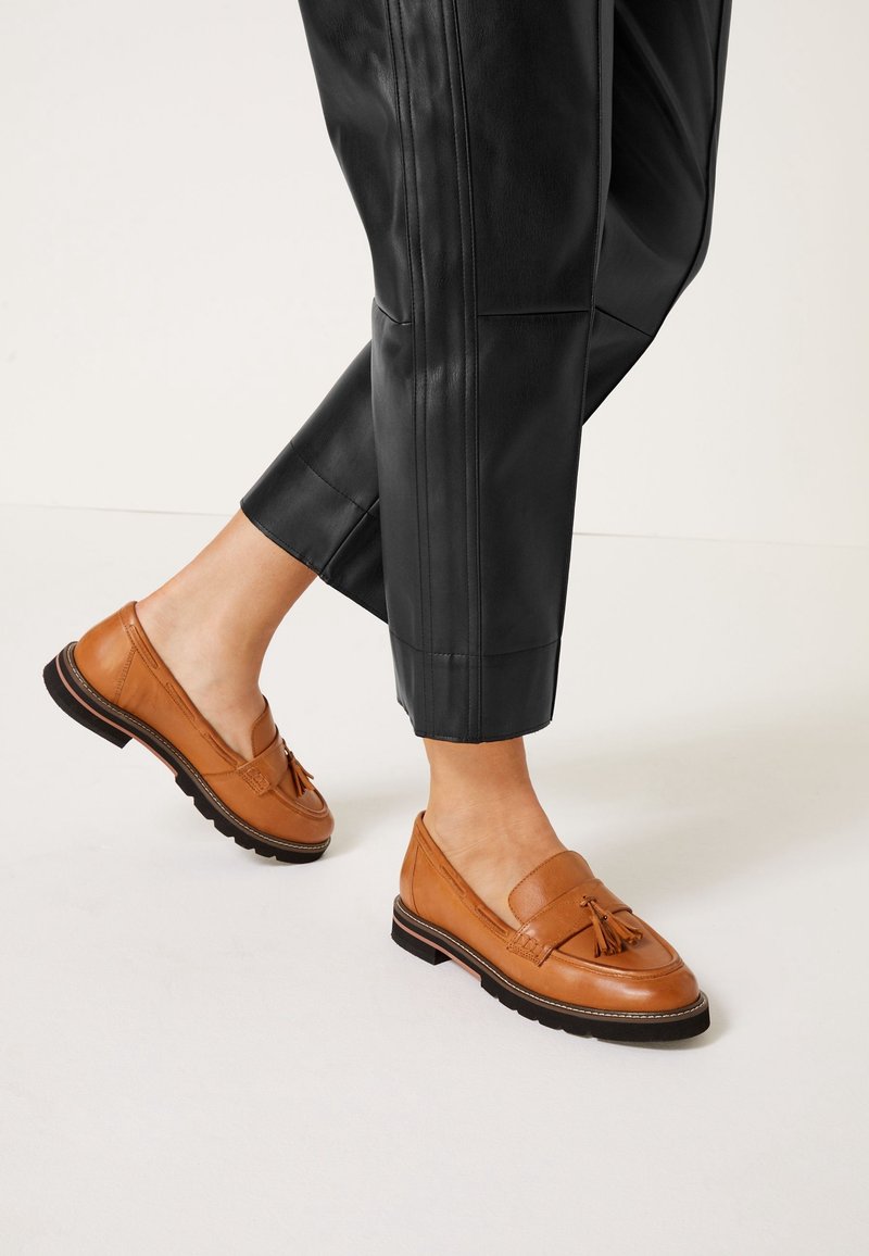 Mocassins en cuir tan avec un détail en pampilles, associés à un pantalon en cuir noir coupé. La chaussure présente une semelle texturée pour une meilleure adhérence.