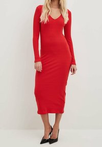 Rotes, geripptes Midi-Kleid mit langen Ärmeln und V-Ausschnitt, figurbetonte Silhouette, glatte Textur, kombiniert mit schwarzen, spitz zulaufenden High Heels.