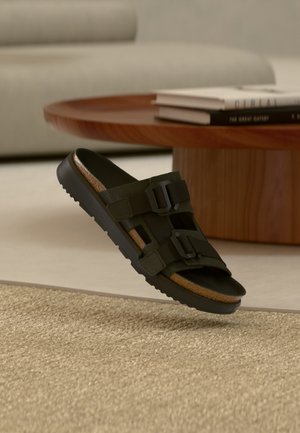 Must avatud varbaga sandaal kahe reguleeritava pandlaga, korkist sisetallaga ja tekstuurse tallaga, asetseb ümmarguse puidust laua lähedal raamatu kuhja kõrval.