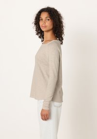 Jauna moteris su garbanotais plaukais, dėvinti smėlio spalvos ilgomis rankovėmis marškinėlį ir šviesiai mėlynas džinsus, stovinti šonu priešais baltą foną.