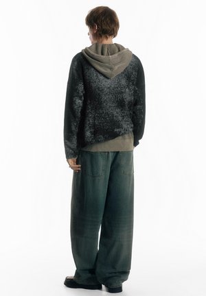 Schwarzer Strickpullover mit strukturiertem Muster, über einem grauen Kapuzenpullover getragen, kombiniert mit lockeren grünen Jeans aus Denim. Outfit von hinten gesehen.