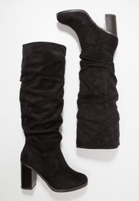 Par de botas altas de ante negro con caña suelta y tacones cuadrados, mostradas planas sobre un fondo blanco.