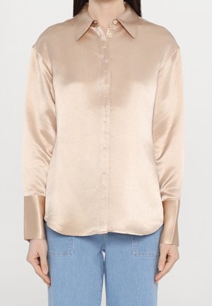 Vrouw draagt een beige satijnen blouse met lange mouwen, brede manchetten en lichtblauwe spijkerbroek, staand tegen een witte achtergrond.
