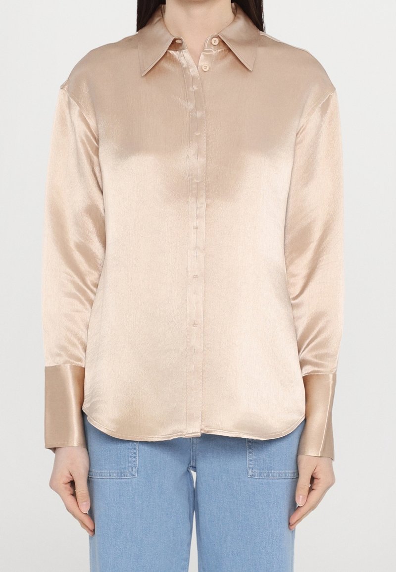 Femme portant une chemise beige en satin à manches longues avec de larges poignets et un jean en denim bleu clair, debout sur un fond blanc.