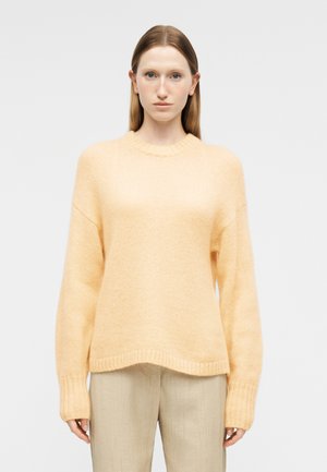 Kvinne med rett hår ikledd en lys gul strikkegenser og lyse beige bukser, stående mot en ensfarget hvit bakgrunn.