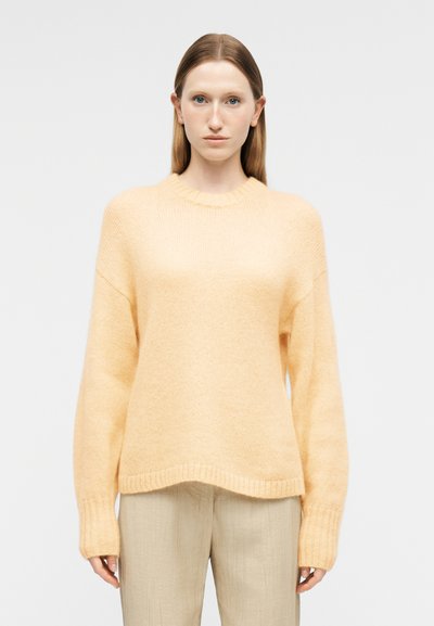 Femme aux cheveux raides portant un pull en maille jaune pâle et un pantalon beige clair, debout devant un fond blanc uni.