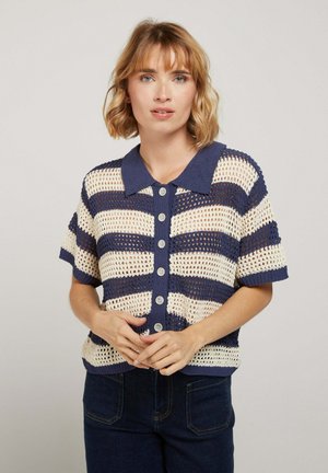 Femme portant un haut à manches courtes, rayé bleu marine et crème, en maille ajourée avec boutons et col, posant devant un fond clair uni.