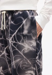 Κοντινό πλάνο μαύρου και άσπρου παντελονιού φόρμας Nike με αφηρημένο καπνιστό σχέδιο, ελαστική μέση με κορδόνι και χέρι στην πλαϊνή τσέπη.