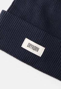 Berretto a maglia a coste blu navy con visiera ripiegata, completo di un'etichetta rettangolare bianca cucita con la scritta "DRYKORN" in lettere nere.