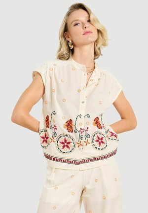 Vrouw met kort blond haar draagt een crèmekleurige blouse met knopen en een broek met oranje bloemendessin, staand met haar handen in de zij.