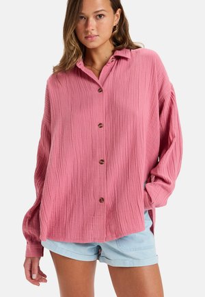 Pink button-up shirt with a textured fabric, long sleeves, and a collar. Features brown buttons and a relaxed fit, paired with light blue shorts.