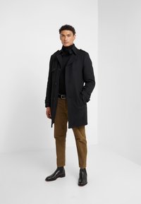 Schwarzer Trenchcoat mit weitem Kragen und Knopfverschluss, kombiniert mit einem schwarzen Rollkragenpullover, hellbraunen Cordhosen und schwarzen Lederstiefeln.