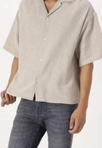 Camicia beige a maniche corte con bottoni e una sottile trama, indossata con jeans neri sbiaditi da una persona davanti a uno sfondo bianco.