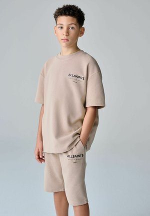 Boy standing with hands in pockets, wearing beige AllSaints short-sleeve top and matching knee-length shorts against plain background.