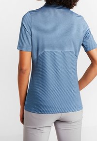 Person klädd i en kortärmad, blårandig polo med ljusgrå byxor, sedd bakifrån mot en enkel vit bakgrund.