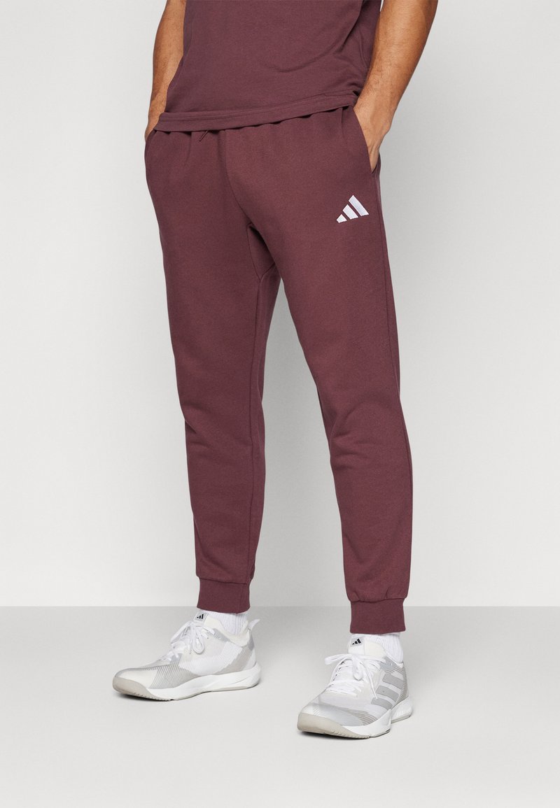 Bordo treniņbikses ar aukliņu jostasvietā, sašaurinātām kājām un baltu Adidas logo kreisajā pusē. Valkātas ar pelēkiem sporta apaviem.