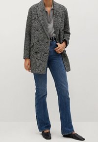 Šedé sako s velkým kopím vzorem a velkými knoflíky, nošené přes šedý top, doplněné modrými rozšířenými džíny a černými mokasíny.