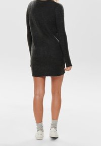 Donna in piedi di spalle, che indossa un vestito a maglia a maniche lunghe grigio scuro, calze grigie e sneakers bianche su uno sfondo grigio.