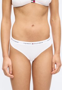 Femme portant un ensemble de sous-vêtements blanc Tommy Hilfiger avec des bandes élastiques à logo, visible de la taille aux cuisses contre un fond uni.