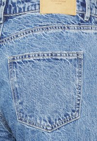 Calças de ganga azul claro com um bolso de remendo na parte de trás, apresentando um acabamento texturizado e detalhes de costura visíveis, com um etiqueta de marca em couro castanho.