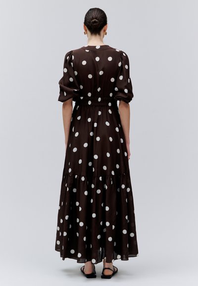 Mujer con cabello recogido en un moño, vestida con un vestido negro con lunares blancos, mangas abullonadas y sandalias, de pie de espaldas sobre un fondo liso.