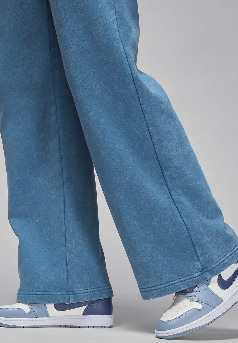 Jambes portant un pantalon large en denim délavé bleu et des baskets blanches avec des accents bleu clair et marine sur un fond gris.