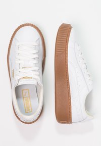 Zapatillas de cuero blancas con suelas de goma marrón texturizadas y punta redonda. Los cordones son blancos y presentan sutiles perforaciones a los lados.