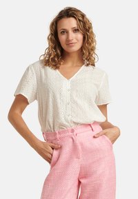 White short-sleeve blouse with eyelet detailing and a V-neck, paired with textured pink high-waisted trousers featuring front pockets.