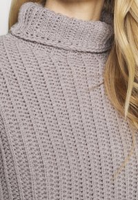 Jasnofioletowy sweter z dzianiny z wysokim, prążkowanym kołnierzem i teksturowanymi liniami pionowymi, prezentujący miękką i przytulną tkaninę.