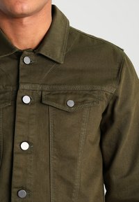 Chaqueta de mezclilla verde oliva con un diseño estructurado, botones de metal plateado, dos bolsillos en el pecho y detalles de costura visibles.