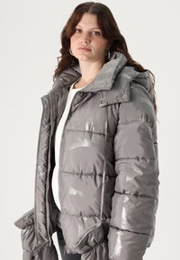 Pufasta jakna od sjajnoga sivog materijala s visokim ovratnikom, zatvaranjem na zatvarač i elastičnim manžetama. Sadrži istaknute šavove i velike džepove.