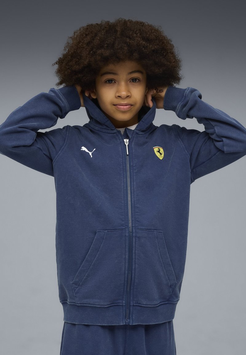 Felpa zip con cappuccio di colore blu navy realizzata in morbido tessuto. Presenta il logo Puma e lo scudo Ferrari sul petto, tasche frontali e polsini a coste.
