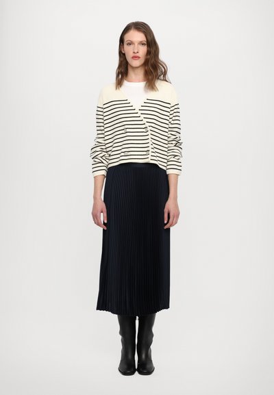 Cardigan crema lavorato a maglia con strisce nere, design doppiopetto; abbinato a una gonna a pieghe blu navy e stivali neri al ginocchio.