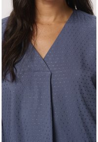 Femme portant un chemisier bleu texturé à col en V, aux longs cheveux foncés reposant sur l'épaule gauche, visible du cou jusqu'au milieu de la poitrine.