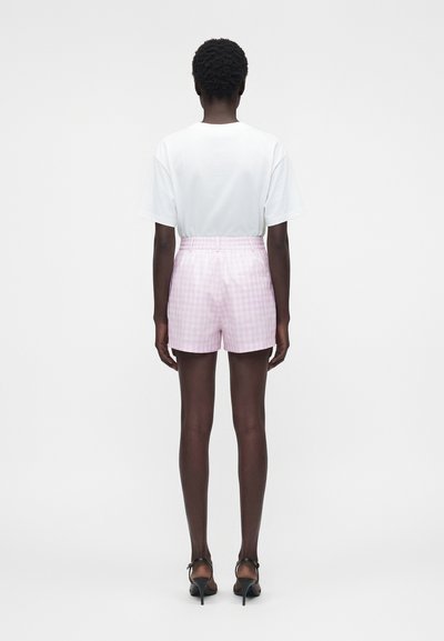 Femme debout de dos, portant un t-shirt blanc, un short à carreaux roses taille haute et des sandales noires à talons sur un fond blanc uni.