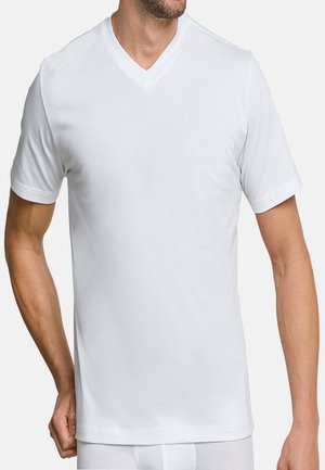 Homme portant un t-shirt blanc à manches courtes et col en V, montré des épaules aux hanches, avec une main ajustant l'ourlet.