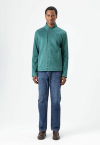 Groene suède jas met een opstaande kraag en een ritssluiting aan de voorkant, gecombineerd met blauwe spijkerbroek en bruine schoenen. De jas heeft een gladde textuur.