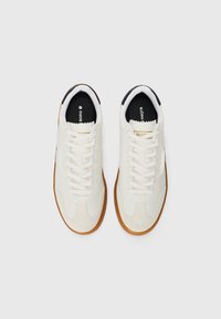 Λευκά αθλητικά παπούτσια με υφή στην άνω επιφάνεια, λείο υλικό, καουτσούκ σόλα, στρογγυλά κορδόνια και μαύρες λεπτομέρειες στη γλώσσα. Ορατό το λογότυπο της μάρκας.